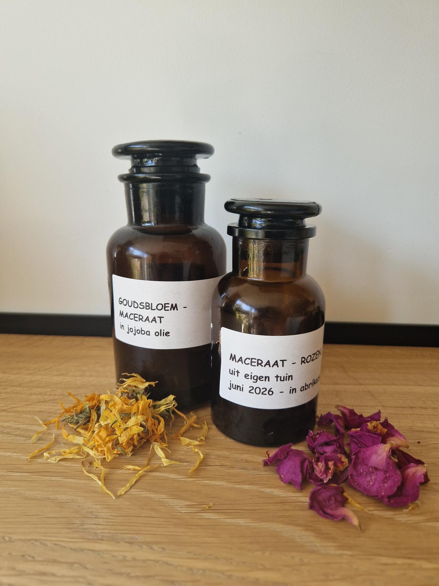 Twee amber glazen potjes met maceraat, met gedroogde goudsbloem en rozenblaadjes op een houten tafel